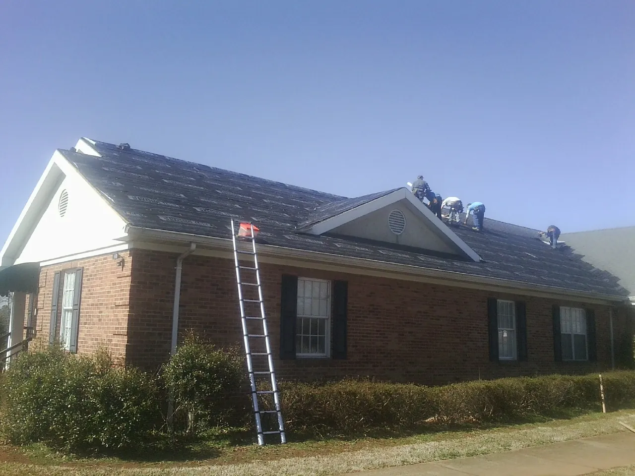 Crew tearing shingles off a brick ranch roof with a ladder set — Metal Roof Repair work by our crew in Harveysburg