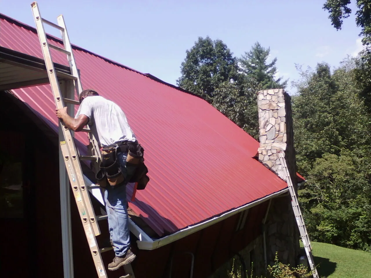 Roofer on a ladder beside a red metal roof and stone chimney — Soffit & Fascia Repair by Roofing Brothers in Harveysburg